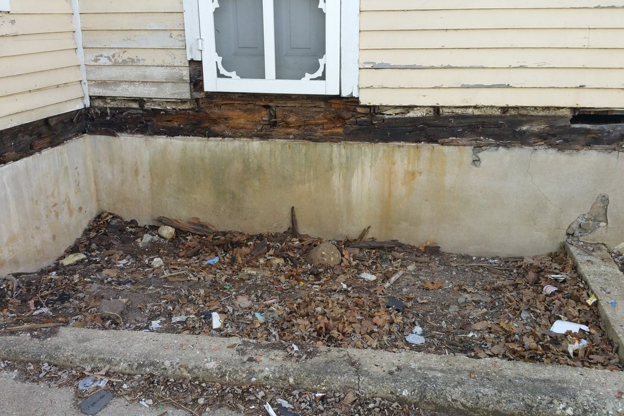 Soil dug back from a house foundation, showing a dark rotted band at the bottom of the siding where dirt sat against it
