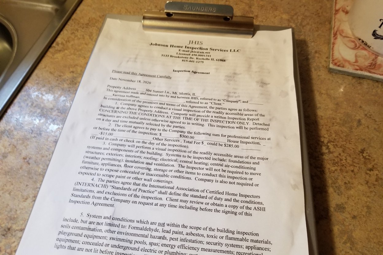A homebuyer reviewing a printed home inspection report while an inspector points to a foundation wall in a basement.