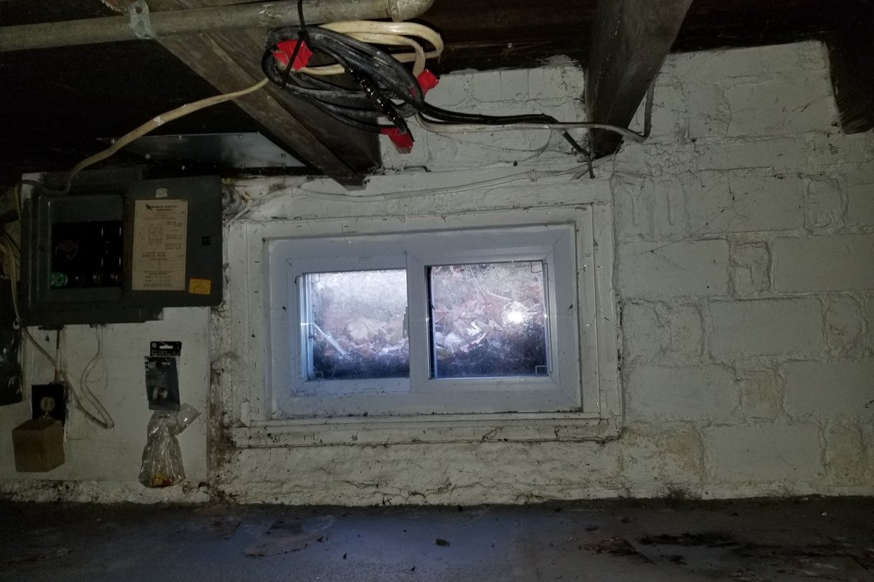 Homeowner with a flashlight inspecting damp wood framing and a plastic ground cover in a dim crawl space.
