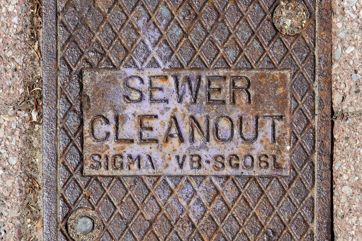 An inspector feeding a sewer camera cable into an open cleanout pipe in a front yard, with the monitor showing the inside of a clay sewer line