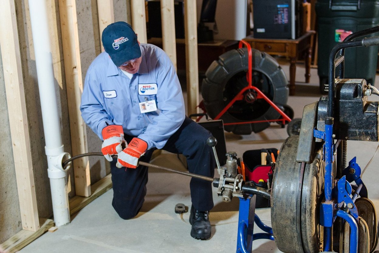 A plumber feeding a sewer camera cable into a cleanout pipe in a basement floor, with a monitor showing the pipe interior