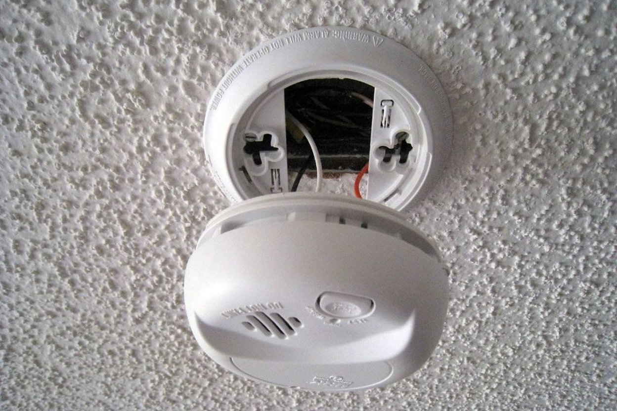 Close-up of a hand pressing the test button on a carbon monoxide alarm mounted on a white wall
