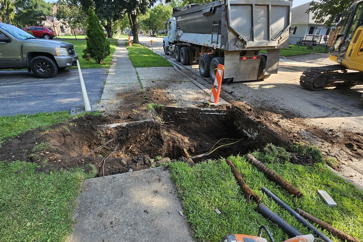 A sewer camera monitor showing a cracked clay pipe, with a plumber feeding the camera cable into a cleanout beside a street curb.