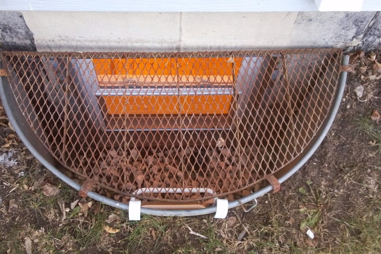 A semicircular basement window well with a metal grate cover and leaves at the bottom